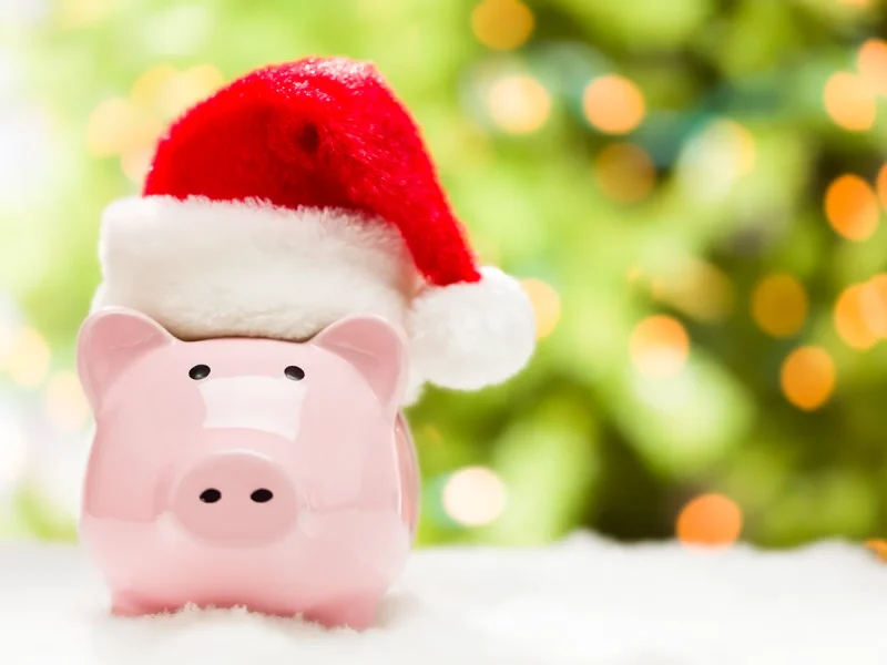 Pink piggy bank wearing a red and white Santa hat. It is standing on on a snowflake covered surface