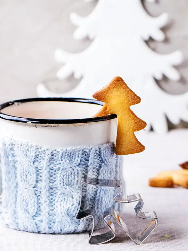 A cup of hot tea in knitted cup holder with a Christmas tree cookie on the side.