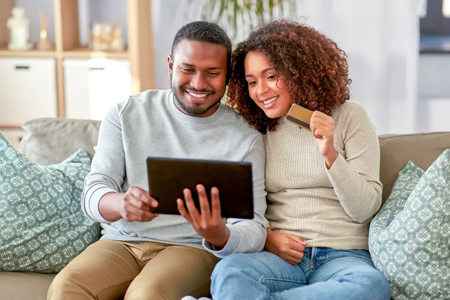 A young couple sitting together on a sofa. They are smiling as they look at a tablet and the woman is holding a credit card ready to go internet shopping