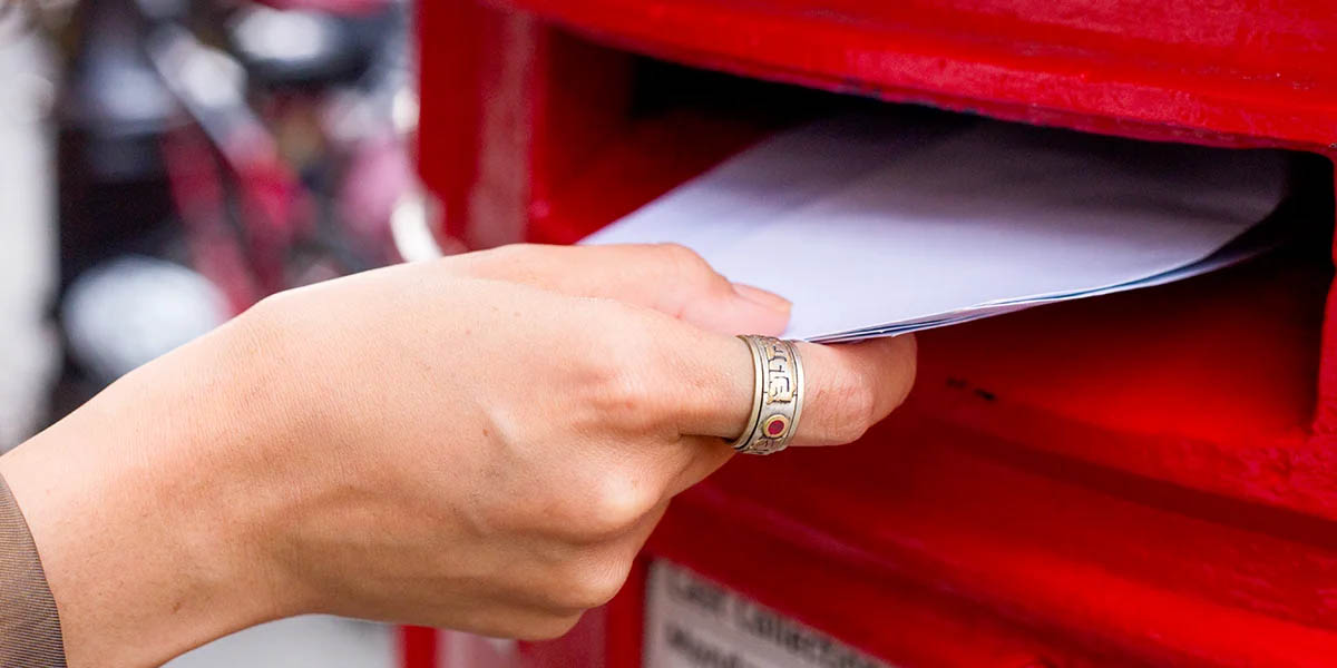 A hand wearing a ring is inserting an envelope into a red mailbox.