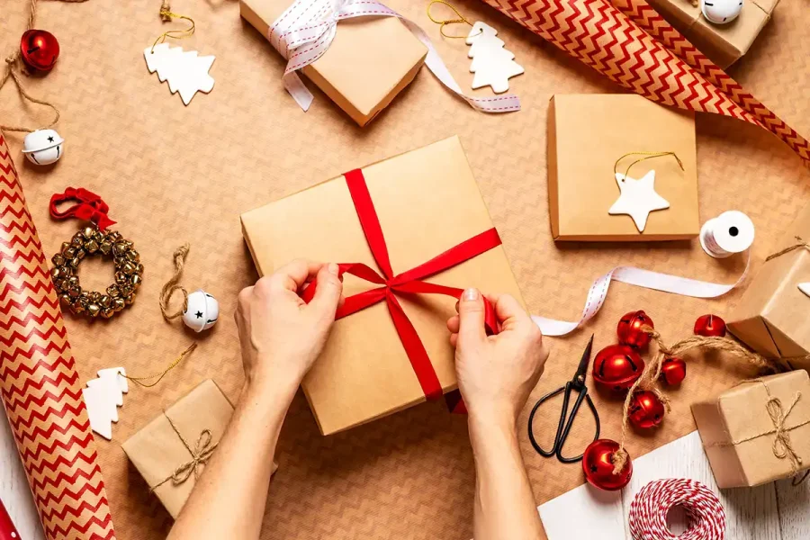 Hands are tying a red ribbon around a gift wrapped in brown paper. The scene shows various wrapped gifts, ornaments, jingle bells, scissors, and rolls of patterned wrapping paper on a table. The background is decorated with festive items for holiday gift wrapping.