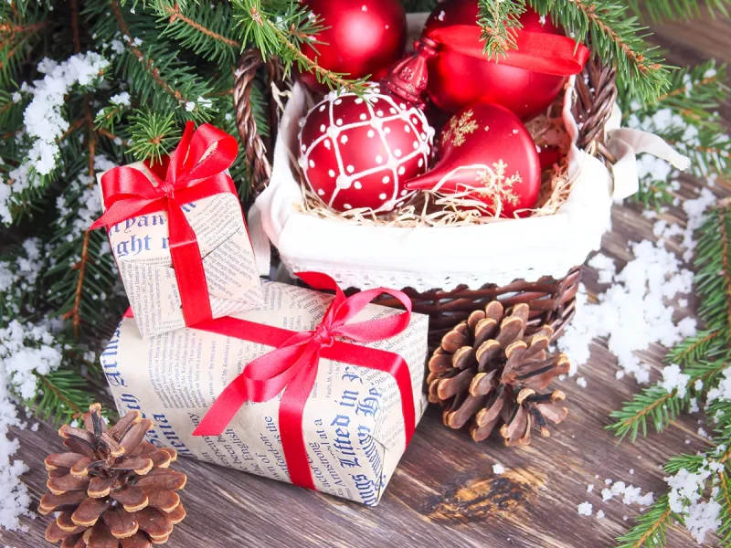 A festive scene featuring red Christmas ornaments in a wicker basket, two wrapped gifts with red ribbons, pine cones, and snow-like decor on a wooden surface, bordered by evergreen branches.