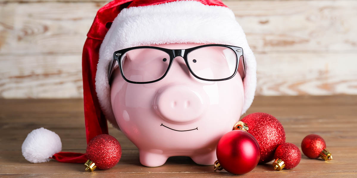 A pink piggy bank wearing black glasses and a red and white Santa hat sits on a wooden surface with red Christmas ornaments scattered around it. The background is a light-colored wooden texture.