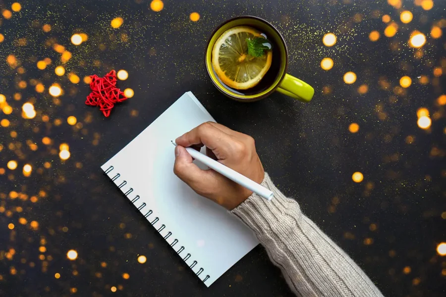 A person's hand holding a pen is poised over an open spiral notebook on a dark surface. A green cup of tea with lemon slices and mint rests beside the notebook. There is a small red star ornament nearby, and golden bokeh lights are scattered in the background.