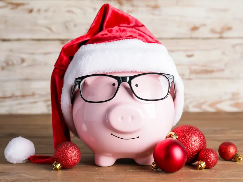A pink piggy bank wearing a red and white Santa hat and black glasses is placed on a wooden surface. Surrounding the piggy bank are red and gold Christmas ornaments. The background consists of wooden planks.