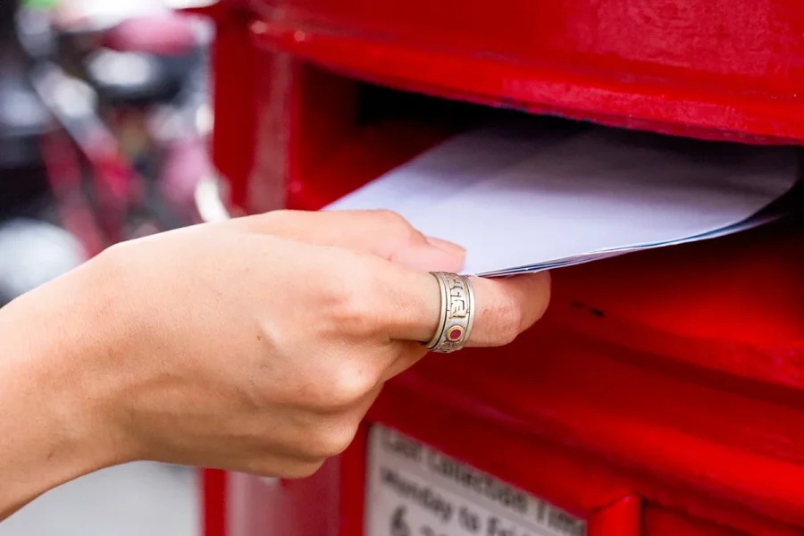 A hand wearing a ring is inserting an envelope into a red postbox. The focus is on the hand and the envelope, with the rest of the background slightly blurred.