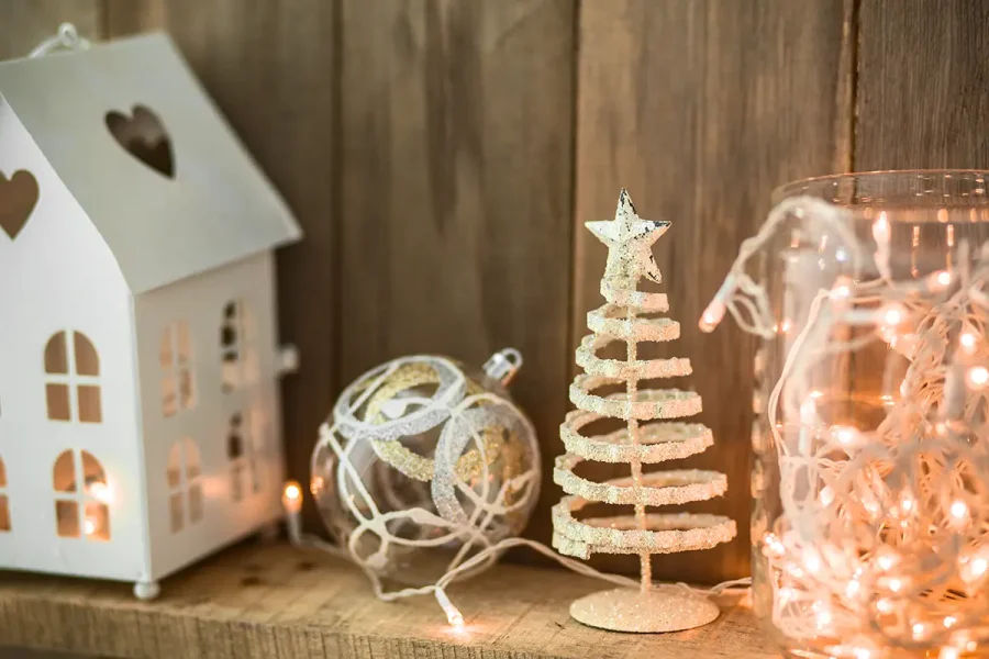 A decorative scene with a white lantern shaped like a house, a spiral Christmas tree figurine with a star on top, a clear jar filled with string lights, and a glittery ornament, all arranged on a wooden surface against a wooden background.