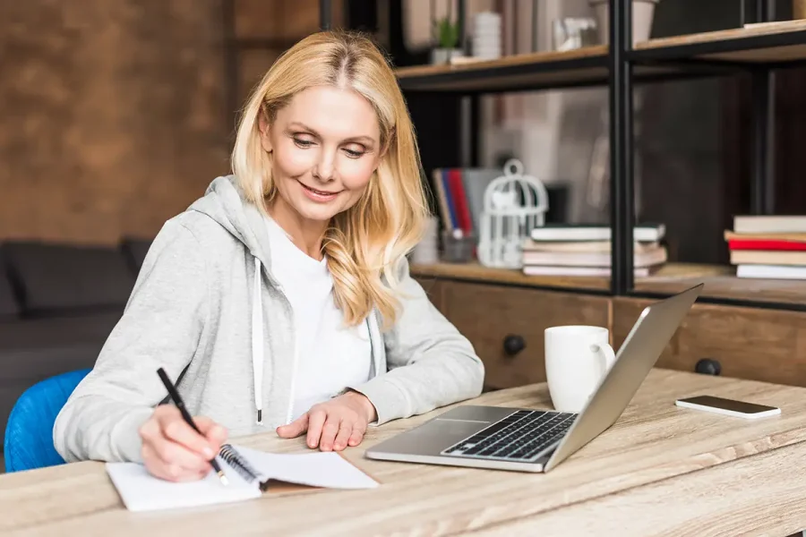 A middle-aged woman sitting at a table, looking at a laptop and making notes in a notepad