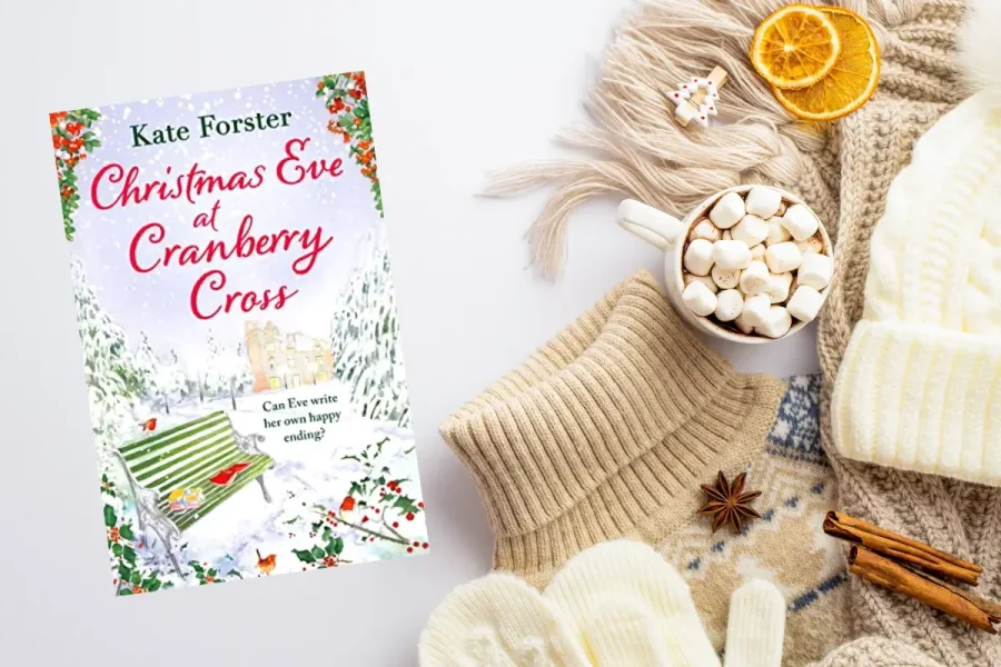 A holiday-themed flat lay image shows a book titled "Christmas Eve at Cranberry Cross" by Kate Forster, with cozy winter items such as cream-colored knitted gloves, a hat, a sweater, a mug of hot chocolate with marshmallows, dried citrus slices, star anise, and cinnamon sticks.
