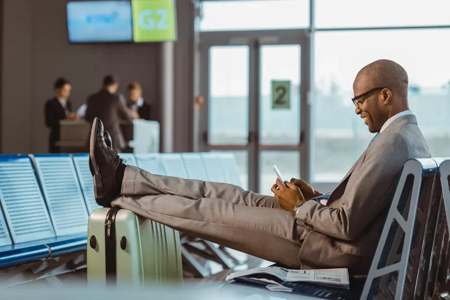 A Black business man smiles confidently as he looks at his mobile phone. He is sitting with his feet propped on his suitcase in an airport departure lounge.