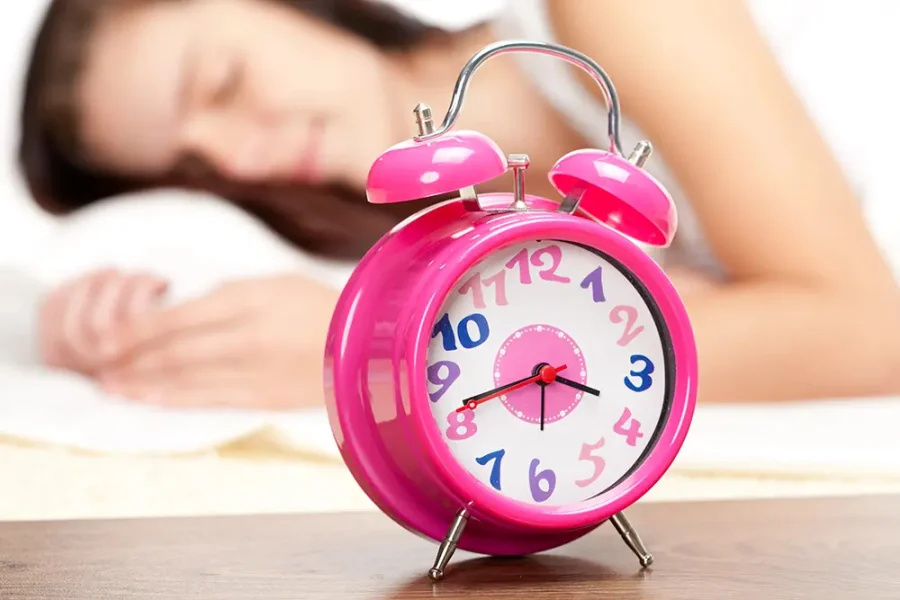 Close up of a pink alarm clock. The blurred image of a woman sleeping can be seen in the background.